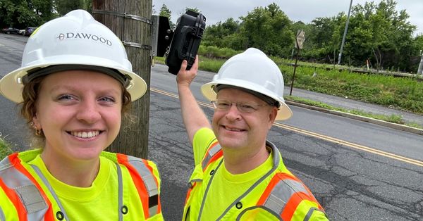Maysen Fisher and Scott Miller wear hard hats and high-visibility safety vests while conducting fieldwork for Dawood Engineering. Maysen smiles at the camera, while Scott holds up field equipment beside a utility pole near a two-lane road.